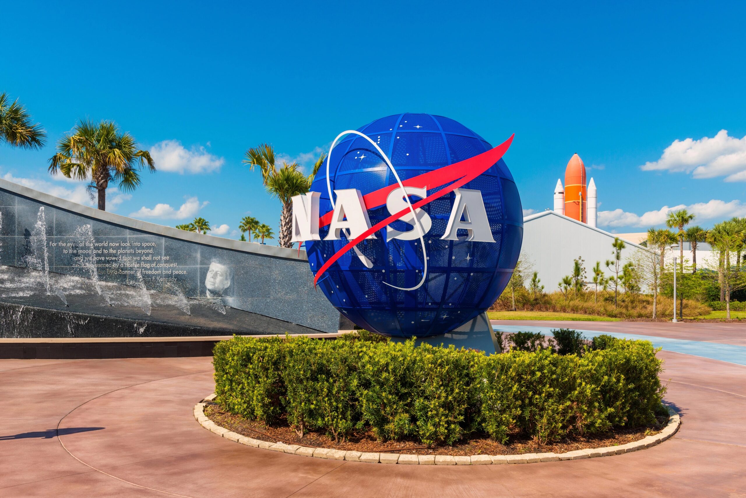 NASA logo sign at the entrance to Kennedy Space Center surrounded by palm trees under a blue Florida sky