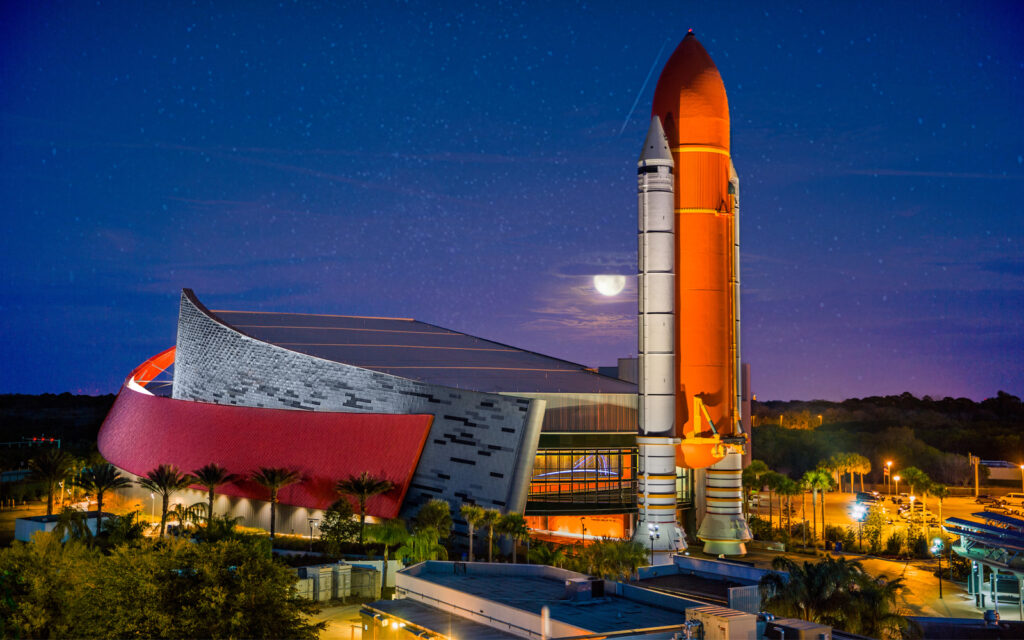 Space shuttle displayed upright at Kennedy Space Center at night with illuminated launch tower and dramatic sky