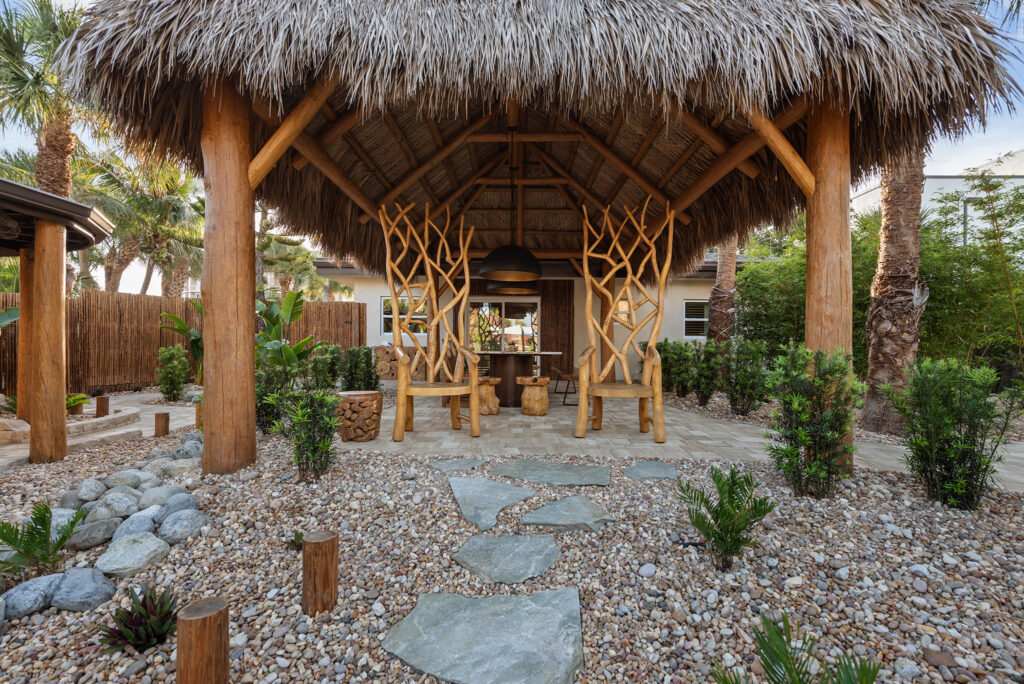 Open air tiki bar with wooden seating, thatched roof, and tropical landscaping