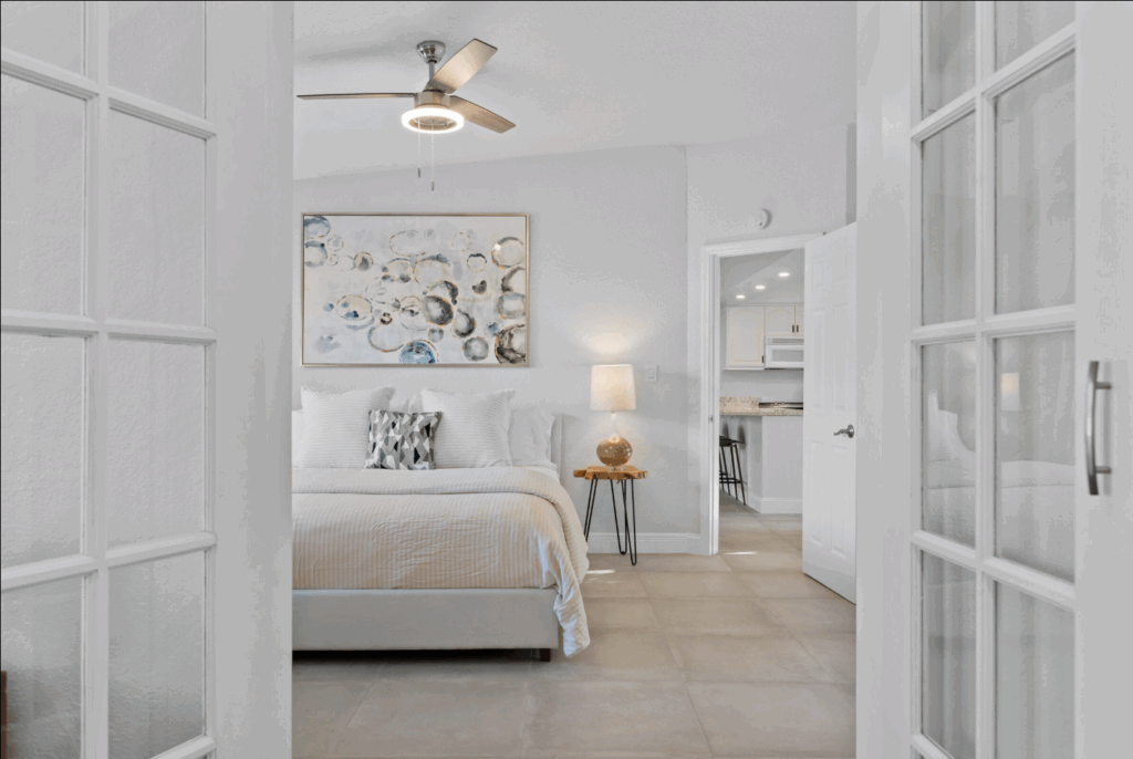 Modern bedroom with white bedding, abstract wall art, and glass French doors opening to a bright, airy interior.