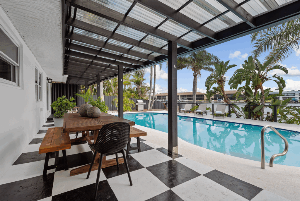 Covered patio with wooden dining table overlooking a modern pool surrounded by palm trees and lounge chairs.