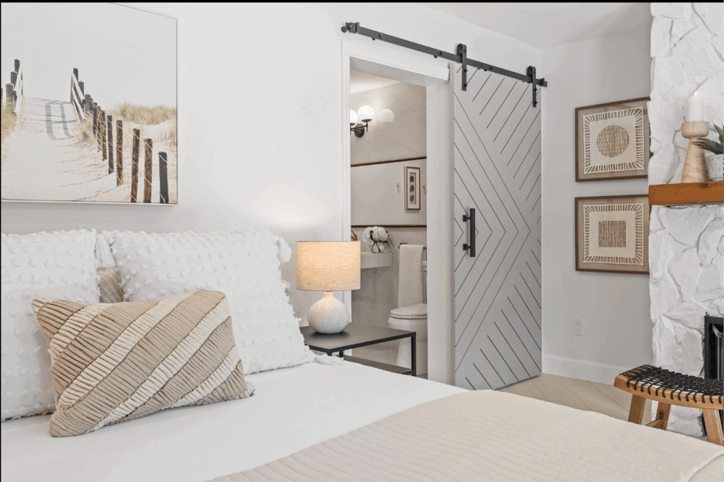 Bright bedroom with white bedding, neutral accent pillows, a barn-style sliding door, and coastal-inspired wall art.